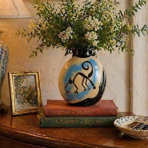 May include: An oval-shaped ceramic vase with a black rim and base, featuring a blue and beige design with a black monkey illustration. The vase holds a bouquet of greenery and white flowers. Two books with titles are stacked below the vase.