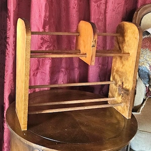 May include: A wooden yarn or thread holder with a natural finish. The rack has two upright sides connected by several horizontal dowels. A central wooden piece is attached to the dowels. The rack is sitting on a dark wooden table.