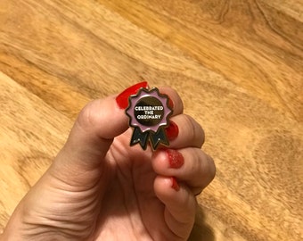 Op de afbeelding: Gouden emaille pin met een roze lint en de tekst "Celebrated the Ordinary".