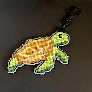 May include: A cross-stitch turtle keychain. The turtle has a green body and a yellow and orange shell. It is attached to a black metal clip. The keychain is a handmade item.