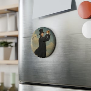 Puede incluir: Un imán redondo con una ilustración de Abraham Lincoln con traje negro y sombrero de copa, jugando al golf. El fondo representa un cielo nublado. El imán está adherido a un refrigerador plateado.