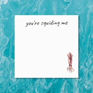 以下が含まれることがあります： 上部に黒の手書き文字で you're squiding me と書かれ、右下隅にピンクがかった茶色のイカの小さな詳細なイラストが描かれた、正方形の白いメモ帳。