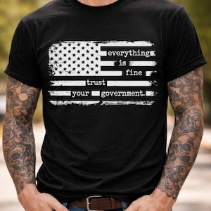 Puede incluir: Camiseta negra con un diseño desgastado de la bandera estadounidense y el texto blanco "everything is fine, trust your government". La camiseta tiene cuello redondo y mangas cortas.