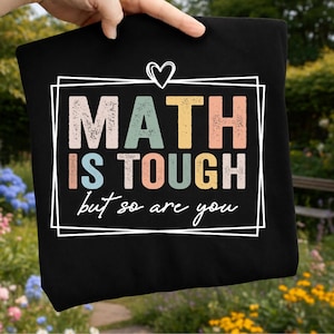 May include: A black t-shirt featuring a white rectangular frame with a heart icon above it. The text "MATH IS TOUGH" is printed in large, distressed, multi-colored block letters, followed by the cursive white text "but so are you".