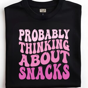 Pode incluir: T-shirt preto de gola redonda com a frase "PROBABLY THINKING ABOUT SNACKS" em rosa, com letras em estilo retro. A camisa tem uma etiqueta na gola. Uma peça de vestuário casual e confortável.