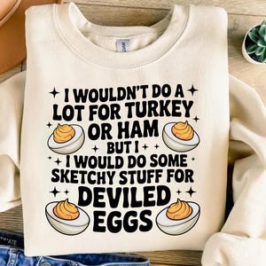 Puede incluir: Sudadera crema con el texto "I WOULDN'T DO A LOT FOR TURKEY OR HAM BUT I WOULD DO SOME SKETCHY STUFF FOR DEVILED EGGS." El diseño incluye ilustraciones de huevos rellenos.