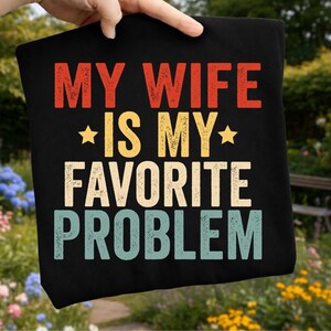 May include: A black t-shirt featuring the text "MY WIFE IS MY FAVORITE PROBLEM" in a distressed, multi-colored font. The words "MY WIFE" are in red, "IS MY" is in yellow with two stars, "FAVORITE" is in cream, and "PROBLEM" is in light blue.