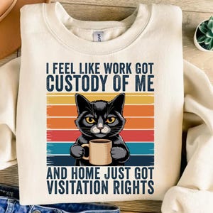 May include: Cream-colored sweatshirt with the text "I FEEL LIKE WORK GOT CUSTODY OF ME AND HOME JUST GOT VISITATION RIGHTS." A black cat with yellow eyes holds a coffee mug against a retro sunset background.