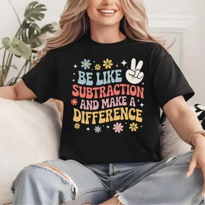 Peut inclure: T-shirt noir avec l'inscription "BE LIKE SUBTRACTION AND MAKE A DIFFERENCE" dans un style rétro coloré. Le design comprend des fleurs et un signe de paix. Un vêtement décontracté pour tous les jours.