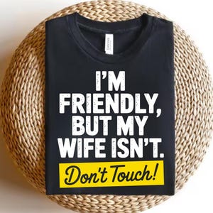 May include: Black t-shirt with the text "I'M FRIENDLY, BUT MY WIFE ISN'T. Don't Touch!" in white and yellow. The shirt is displayed on a woven, circular surface.