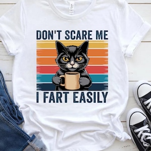 May include: White t-shirt featuring a black cat holding a coffee mug. The shirt has the text "DON'T SCARE ME I FART EASILY" above and below the cat graphic, set against a retro sunset-style background.