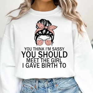 以下が含まれることがあります： 「YOU THINK I'M SASSY YOU SHOULD MEET THE GIRL I GAVE BIRTH TO」のテキストが入った白いスウェットシャツ。グラフィックは、お団子ヘア、サングラス、ヒョウ柄のヘッドバンドをした女性の顔が特徴です。