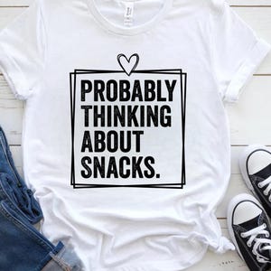 Pode incluir: Camiseta branca com a frase "PROBABLY THINKING ABOUT SNACKS" em negrito preto, emoldurada por um quadrado preto e um coração. A camiseta está dobrada, com tênis pretos e brancos visíveis no canto.