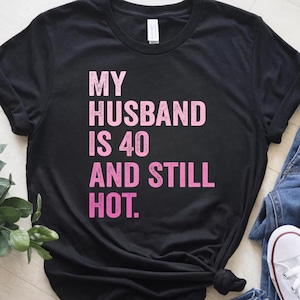 May include: Black t-shirt with the text "MY HUSBAND IS 40 AND STILL HOT" in pink gradient lettering. The shirt is a crew neck style and appears to be made of a soft material. The shirt is styled with blue jeans and white sneakers.