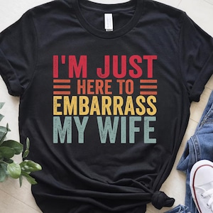 Puede incluir: Camiseta negra con el texto "I'M JUST HERE TO EMBARRASS MY WIFE" en letras rojas, naranjas, amarillas y verde azulado. La camiseta está hecha de un material suave y tiene un cuello redondo clásico.