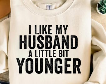Könnte beinhalten: Cremefarbenes Sweatshirt mit dem Text "I LIKE MY HUSBAND A LITTLE BIT YOUNGER" in fetten schwarzen Buchstaben. Das Sweatshirt ist gefaltet, mit einem Blick auf Jeans am unteren Rand. Eine kleine Sukkulentenpflanze ist im Hintergrund.