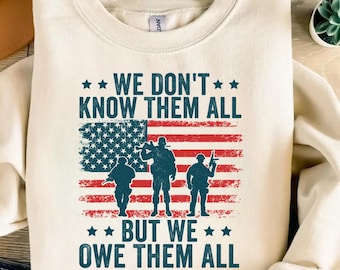 May include: Cream-colored sweatshirt with the text "WE DON'T KNOW THEM ALL BUT WE OWE THEM ALL" in blue. The design includes an American flag and silhouettes of three soldiers. The sweatshirt is a crew neck style.