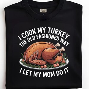 以下が含まれることがあります： 黒いスウェットシャツ。白いお皿に載ったローストターキーのグラフィック、緑と赤いベリーに囲まれています。「I COOK MY TURKEY THE OLD FASHIONED WAY I LET MY MOM DO IT」というテキストが入っています。