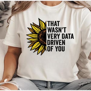 Pode incluir: Camiseta branca com um girassol e o texto "THAT WASN'T VERY DATA DRIVEN OF YOU" em preto. O girassol tem pétalas amarelas e um centro castanho.