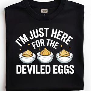 Può includere: T-shirt nero girocollo con la scritta bianca "I'M JUST HERE FOR THE DEVILED EGGS". Il design include tre uova alla diavola con ripieno giallo. Una divertente maglietta grafica a tema cibo.
