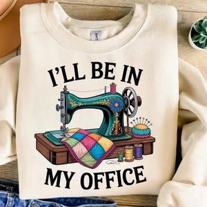 Puede incluir: Sudadera color crema con el texto "I'LL BE IN MY OFFICE." El gráfico presenta una máquina de coser turquesa, un edredón colorido, carretes de hilo y un alfiletero. Ideal para los entusiastas de la costura.