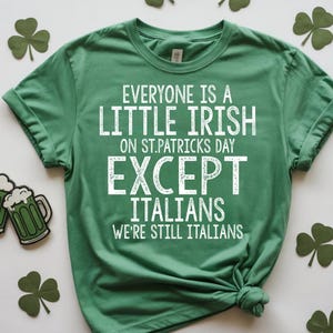 以下が含まれることがあります： 白文字で「EVERYONE IS A LITTLE IRISH ON ST.PATRICKS DAY EXCEPT ITALIANS WE'RE STILL ITALIANS」と書かれた緑色のTシャツ。シャツは裾で結ばれています。背景にはクローバーとビールジョッキの装飾があります。