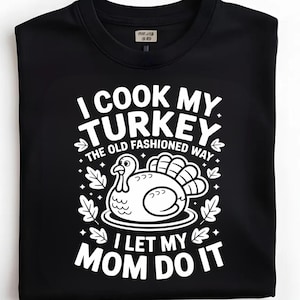 以下が含まれることがあります： 黒いクルーネックTシャツ。白いグラフィックには「I COOK MY TURKEY THE OLD FASHIONED WAY I LET MY MOM DO IT」と書かれており、漫画の七面鳥と葉のアクセントが描かれています。
