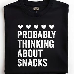 Pode incluir: Camisola preta de gola redonda com texto branco e gráficos de coração. O texto diz "PROBABLY THINKING ABOUT SNACKS" em negrito. Acima do texto, cinco símbolos de coração brancos. A camisola tem uma etiqueta na gola.