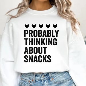 Pode incluir: Camisola branca com o texto "PROBABLY THINKING ABOUT SNACKS" em letras pretas em negrito. Acima do texto estão cinco símbolos de coração preto. A camisola é combinada com jeans azuis.
