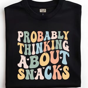 Pode incluir: Camisola preta de gola redonda com o texto "PROBABLY THINKING ABOUT SNACKS" em uma fonte retrô e colorida. O texto é em tons de laranja, amarelo, azul e verde. A camisola tem uma etiqueta na gola.