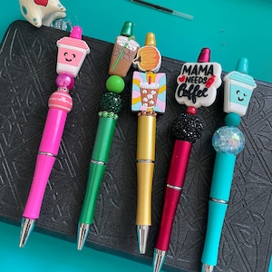 May include: Five pens with decorative toppers and beads. The pens are pink, green, gold, red, and teal. Toppers include a pink cup, iced coffee, a pumpkin, a sign reading MAMA NEEDS Coffee, and a teal cup. A small white cat figurine is visible.
