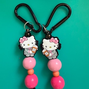 May include: Two identical keychains featuring a white Hello Kitty character wearing a gray suit and holding an orange prescription bottle. Each keychain includes a black carabiner, a dark bead, a large pink bead, a small orange bead, and a large hot pink bead.