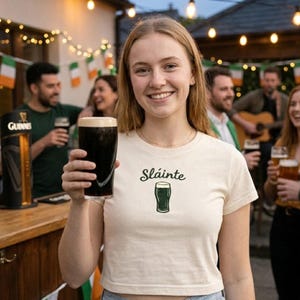Könnte beinhalten: Eine Frau hält ein Pint dunkles Bier und trägt ein cremefarbenes T-Shirt mit dem Wort "Sláinte" und einer Bierglas-Grafik. Im Hintergrund sind eine Bar, irische Flaggen und Menschen beim Trinken zu sehen.