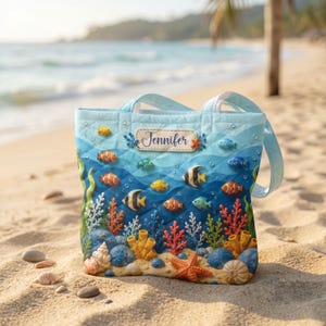 May include: A light blue tote bag with a colourful underwater scene featuring various fish, coral, and sea life. The bag has a quilted design and the name "Jennifer" is displayed on a label. The bag has light blue handles.