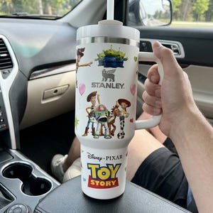 May include: White Stanley tumbler featuring Toy Story characters, including Woody, Buzz Lightyear, and the Little Green Men. The tumbler has a handle, a straw, and the Disney Pixar Toy Story logo. The tumbler is in a car.