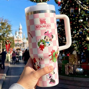May include: A pink and white checkered Stanley tumbler featuring Minnie Mouse and holiday-themed graphics. The tumbler has a white handle and lid, with the Stanley logo visible. The design includes images of Minnie Mouse with a wreath, gifts, and candy canes.