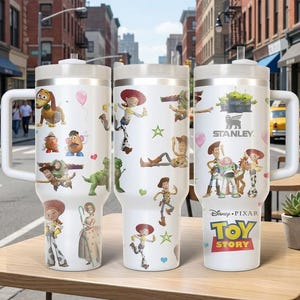 May include: Three white Stanley tumblers featuring characters from Disney Pixar's Toy Story. Each tumbler has a white handle and lid, with colorful graphics of Woody, Buzz Lightyear, Jessie, and other characters. The tumblers are on a wooden table.