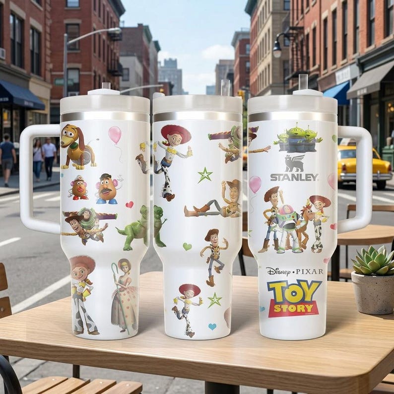 May include: Three white Stanley tumblers featuring characters from Disney Pixar's Toy Story. Each tumbler has a white handle and lid, with colorful graphics of Woody, Buzz Lightyear, Jessie, and other characters. The tumblers are on a wooden table.