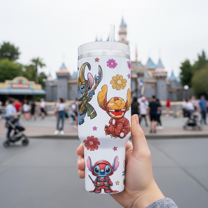 May include: White tumbler featuring cartoon characters and floral designs. The tumbler has a clear lid and a straw. The characters are in various costumes. The background shows a theme park with a castle.
