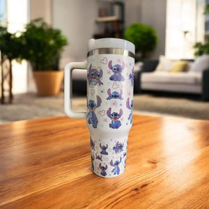 K&ouml;nnte beinhalten: Ein wei&szlig;er Reisebecher mit Henkel, verziert mit einem sich wiederholenden Muster der Figur Stitch in Blau mit rosa Akzenten. Das Design zeigt Stitch in verschiedenen Posen, vor einem Hintergrund aus kleinen rosa Blumen und Sternen. Der Becher hat einen wei&szlig;en Deckel.