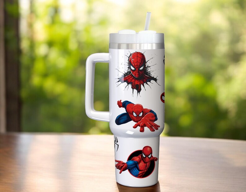 Puede incluir: Vaso t&eacute;rmico blanco con gr&aacute;ficos de Spider-Man. El vaso tiene un asa, una tapa y una pajita. Los gr&aacute;ficos de Spider-Man son en rojo, azul y negro. El vaso est&aacute; sobre una superficie de madera.