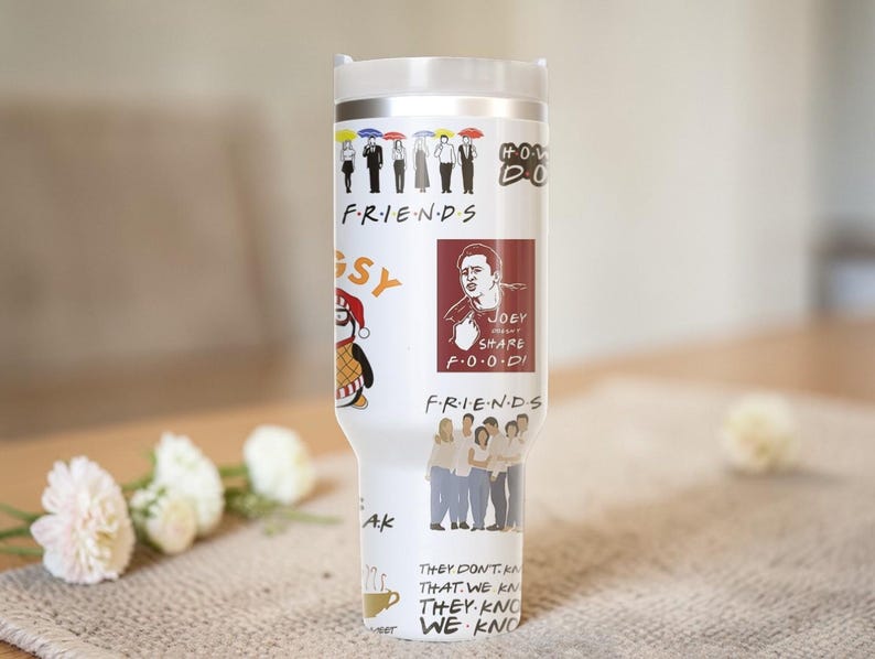 Puede incluir: Un vaso de acero inoxidable blanco con varios gr&aacute;ficos y texto de la serie de televisi&oacute;n "Friends". El dise&ntilde;o incluye personajes de dibujos animados, el logotipo del programa y citas. El vaso tiene una tapa blanca y un borde plateado.