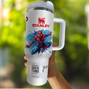 Puede incluir: Vaso Stanley Quencher H2.0 Flowstate blanco con un gr&aacute;fico de Spider-Man. El vaso presenta un dise&ntilde;o de Spider-Man rojo y azul sobre un fondo de telara&ntilde;a azul. Tiene un asa y tapa blancos, y el logotipo de Stanley es visible.