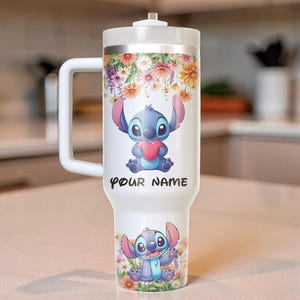 Puede incluir: Un vaso t&eacute;rmico blanco con asa, con un dise&ntilde;o floral y un personaje de dibujos animados sosteniendo un coraz&oacute;n. El texto "YOUR NAME" est&aacute; impreso en el vaso. El vaso tiene una capacidad aproximada de 887 ml.