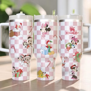 May include: Three stainless steel tumblers with white handles and lids. The tumblers have a pink and white checkered pattern with Disney holiday characters and candy cane designs. The Stanley logo is visible on one tumbler.