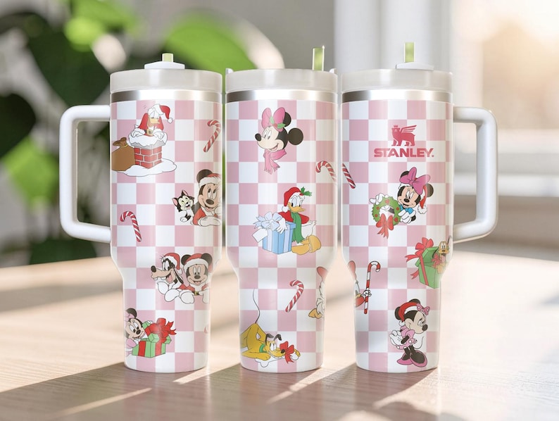 May include: Three stainless steel tumblers with white handles and lids. The tumblers have a pink and white checkered pattern with Disney holiday characters and candy cane designs. The Stanley logo is visible on one tumbler.