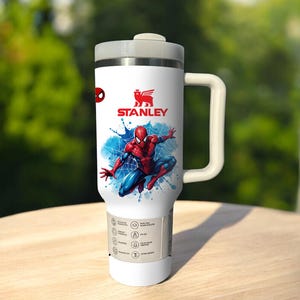 Puede incluir: Taza de viaje Stanley blanca con un dise&ntilde;o de Spider-Man. La taza tiene una tapa y un asa grises, con el logotipo de Stanley y una ilustraci&oacute;n de Spider-Man en rojo y azul. La taza tiene una base blanca y una etiqueta con iconos.