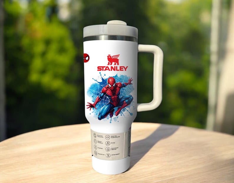 Puede incluir: Taza de viaje Stanley blanca con un dise&ntilde;o de Spider-Man. La taza tiene una tapa y un asa grises, con el logotipo de Stanley y una ilustraci&oacute;n de Spider-Man en rojo y azul. La taza tiene una base blanca y una etiqueta con iconos.
