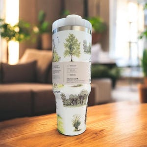 K&ouml;nnte beinhalten: Ein wei&szlig;-silberner Isolierbecher mit einem Design zum Thema Natur. Der Becher zeigt Illustrationen von B&auml;umen und Landschaften. Der Becher hat einen wei&szlig;en Deckel und ein silbernes Band. Der Becher steht auf einer Holzoberfl&auml;che.