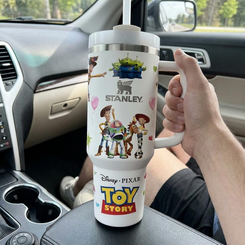 May include: White Stanley tumbler featuring Toy Story characters, including Woody, Buzz Lightyear, and the Little Green Men. The tumbler has a handle, a straw, and the Disney Pixar Toy Story logo. The tumbler is in a car.